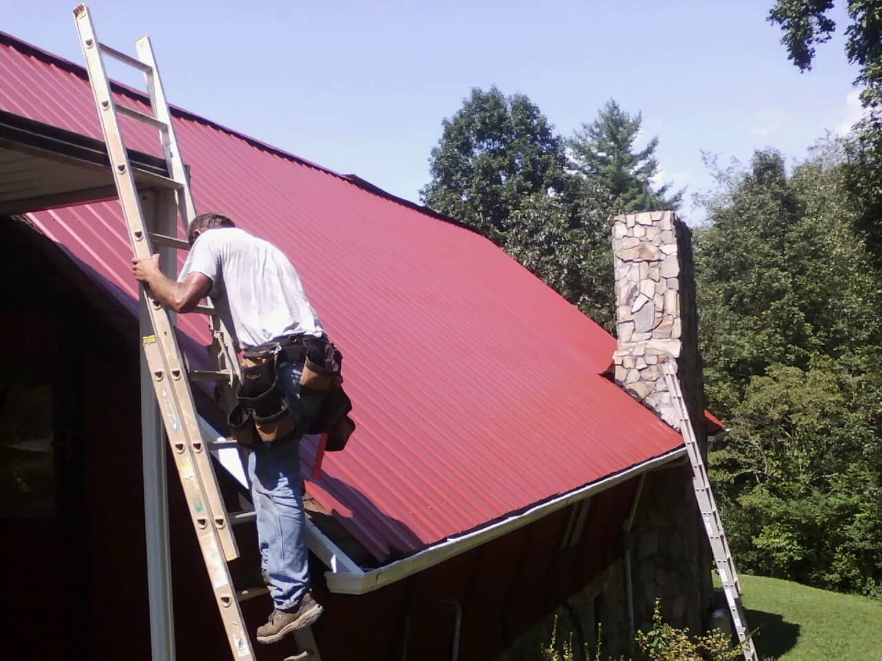 Roofer on a ladder beside a red metal roof and stone chimney — Soffit & Fascia Repair by Roofing Brothers in Holtwood